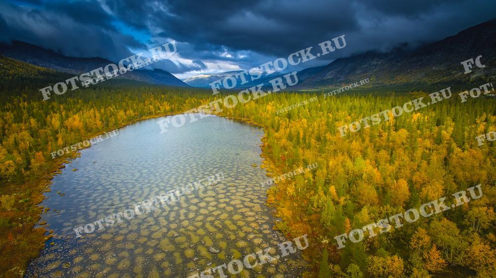 На фотографии изображены Хибины — горный массив в Мурманской области. В центре кадра спокойное озеро, окруженное лесами, чьи деревья окрашены в золотистые осенние оттенки. Тёмные облака придают пейзажу атмосферу суровости северной природы. Хибины — один из крупнейших горных массивов Кольского полуострова, популярный среди любителей активного отдыха и горных походов.